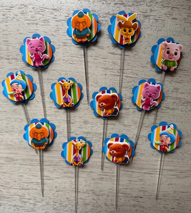 May include: A collection of colorful cupcake toppers featuring various cartoon characters. Each topper has a blue scalloped edge, a striped background, and a character image. The toppers are on clear plastic sticks.
