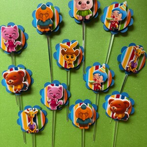 May include: A collection of colorful cupcake toppers featuring various cartoon animal characters. Each topper has a blue scalloped edge, a striped background, and a clear plastic stick. The characters include a pig, cat, bear, giraffe, and elephant.