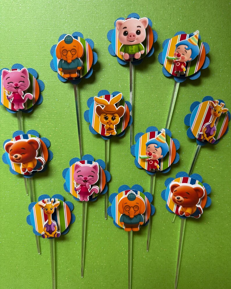 May include: A collection of colorful cupcake toppers featuring various cartoon animal characters. Each topper has a blue scalloped edge, a striped background, and a clear plastic stick. The characters include a pig, cat, bear, giraffe, and elephant.