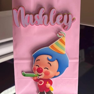 Puede incluir: Una bolsa de regalo rosa con el nombre "Nashley" en letras rosas y doradas. Un payaso de dibujos animados con pelo azul, una nariz roja y un gorro de fiesta est&aacute; en la bolsa. El payaso sostiene un cuerno de fiesta verde.