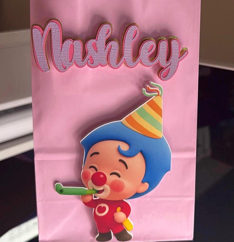 Puede incluir: Una bolsa de regalo rosa con el nombre "Nashley" en letras rosas y doradas. Un payaso de dibujos animados con pelo azul, una nariz roja y un gorro de fiesta est&aacute; en la bolsa. El payaso sostiene un cuerno de fiesta verde.