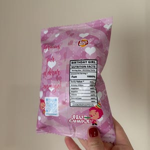 May include: A pink Lay's potato chip bag with a birthday theme. The bag features text in Spanish and English, heart patterns, and a cartoon illustration. The bag is held by a hand with red nail polish.