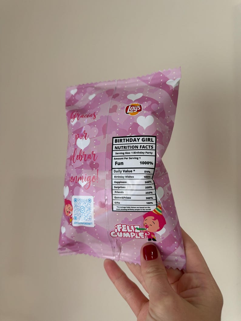 May include: A pink Lay's potato chip bag with a birthday theme. The bag features text in Spanish and English, heart patterns, and a cartoon illustration. The bag is held by a hand with red nail polish.
