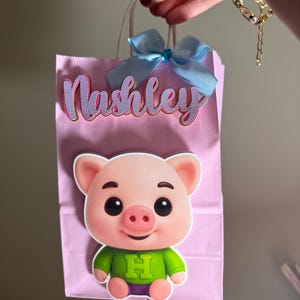Puede incluir: Bolsa de regalo rosa con un lazo azul claro y el nombre "Nashley" en letras moradas y doradas. Un cerdo de dibujos animados con una camisa verde con una gran "H" est&aacute; adherido a la parte delantera de la bolsa.