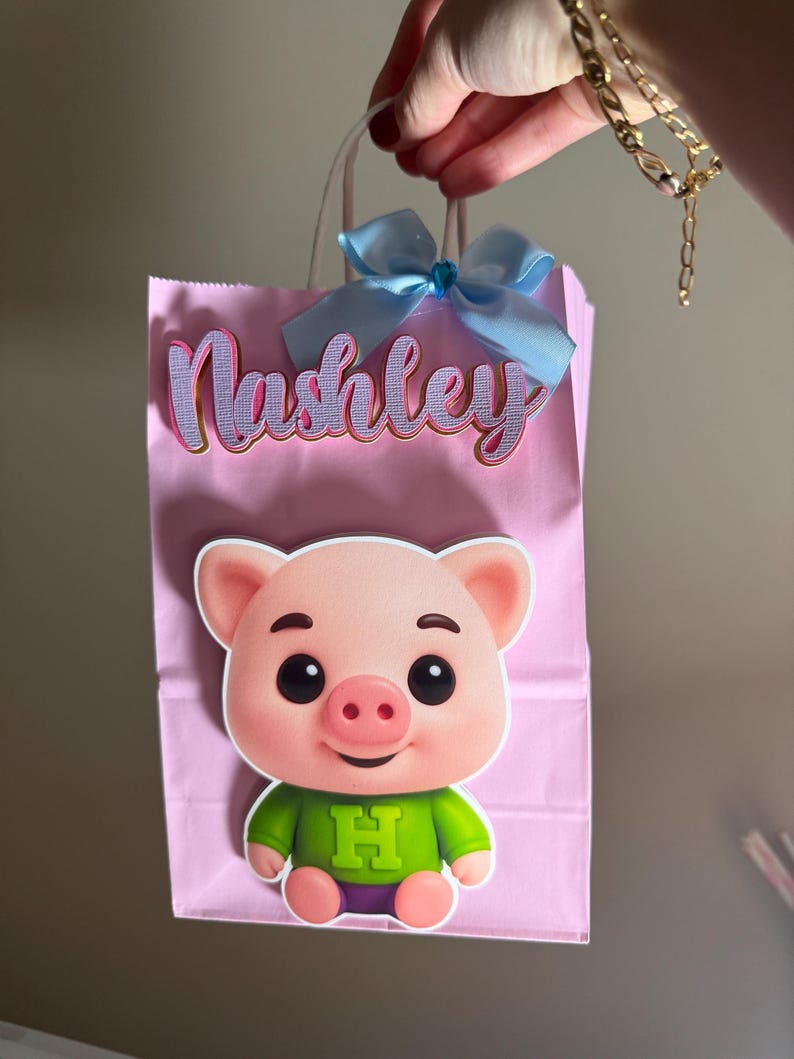Puede incluir: Bolsa de regalo rosa con un lazo azul claro y el nombre "Nashley" en letras moradas y doradas. Un cerdo de dibujos animados con una camisa verde con una gran "H" est&aacute; adherido a la parte delantera de la bolsa.