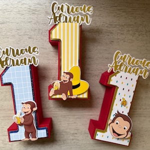 May include: Three red number one decorations with Curious George themes. One has a blue grid pattern, another has yellow stripes, and the third has white polka dots. Each has a Curious George character and the text "Curious Adrian".
