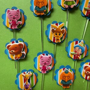 May include: A collection of colorful cupcake toppers featuring cartoon animal characters. Each topper has a blue scalloped edge, a striped background, and a clear plastic stick. The characters include a pig, bear, cat, elephant, and giraffe.
