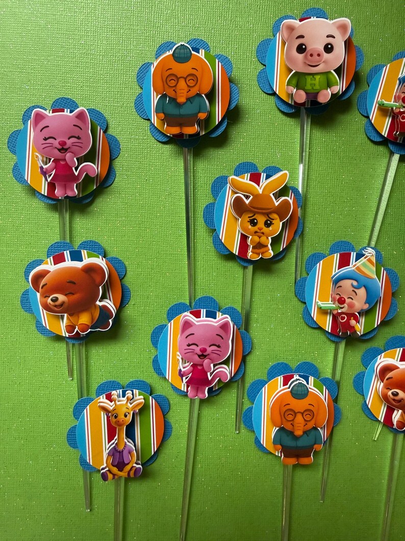 May include: A collection of colorful cupcake toppers featuring cartoon animal characters. Each topper has a blue scalloped edge, a striped background, and a clear plastic stick. The characters include a pig, bear, cat, elephant, and giraffe.