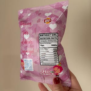 May include: A pink Lay's potato chip bag with a birthday theme. The bag features heart patterns, text in Spanish, and a nutrition facts label. The text on the bag says "Birthday Girl" and "Feliz Cumple".