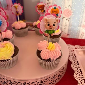 Pu&ograve; includere: Una collezione di cupcakes con glassa rosa e gialla, ognuno sormontato da un personaggio decorativo. I cupcakes sono disposti su un supporto bianco a pi&ugrave; livelli con un bordo decorativo, su una tovaglia rossa.