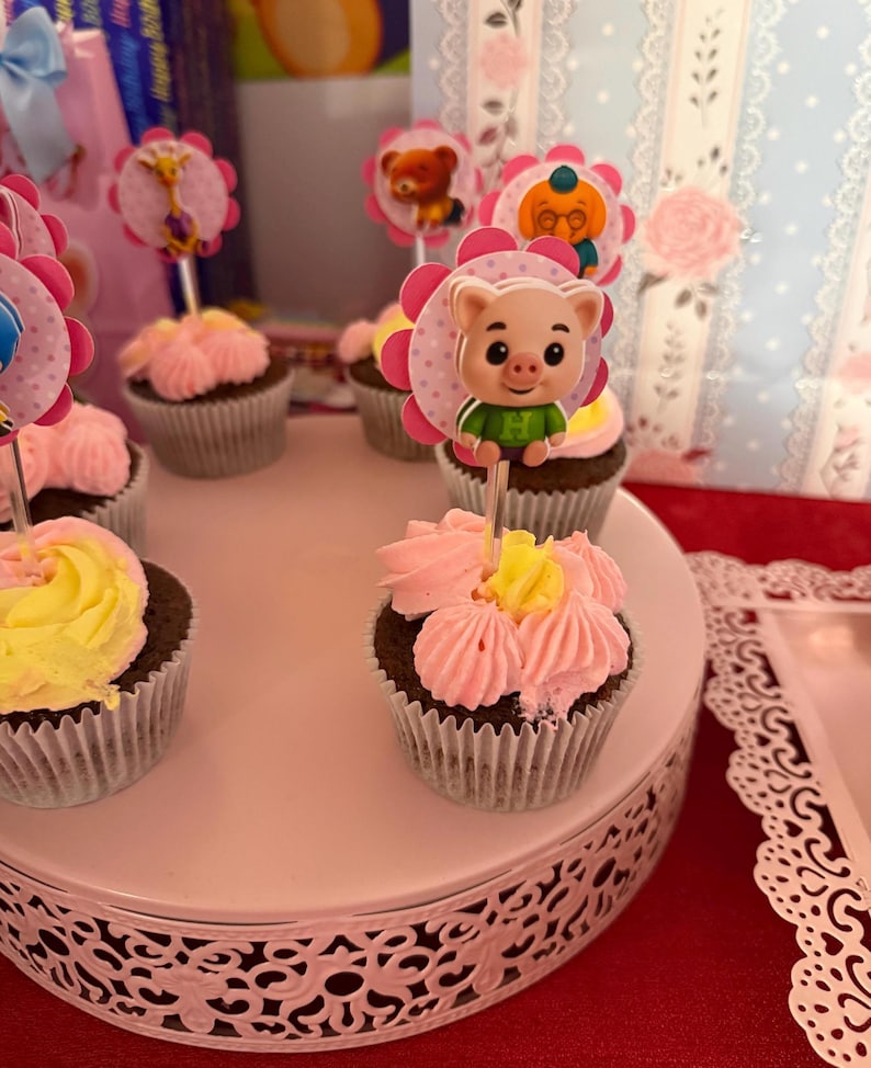 Pu&ograve; includere: Una collezione di cupcakes con glassa rosa e gialla, ognuno sormontato da un personaggio decorativo. I cupcakes sono disposti su un supporto bianco a pi&ugrave; livelli con un bordo decorativo, su una tovaglia rossa.