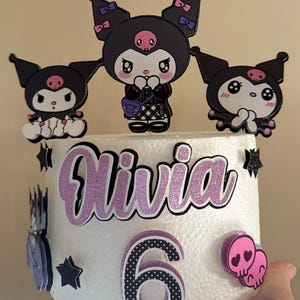 Könnte beinhalten: Eine weiße Torte, verziert mit drei schwarzen Cartoon-Figuren mit rosa Akzenten. Der Kuchen ist mit dem Namen "Olivia" in glitzernden lila Buchstaben und der Zahl "6" verziert. Zusätzliche Dekorationen sind schwarze Sterne und rosa Schädel-Herz-Formen.