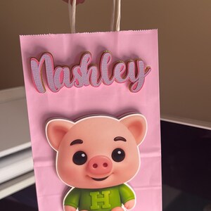 Puede incluir: Bolsa de regalo rosa con una ilustraci&oacute;n de cerdo de dibujos animados y el nombre "Nashley" en cursiva. El cerdo lleva una camisa verde con una "H" morada. La bolsa tiene asas de cuerda blanca.