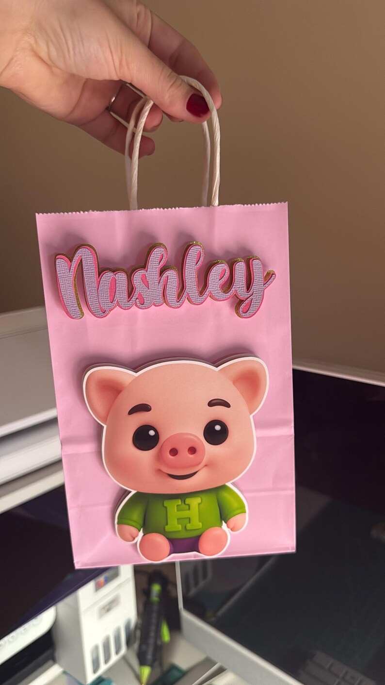 Puede incluir: Bolsa de regalo rosa con una ilustraci&oacute;n de cerdo de dibujos animados y el nombre "Nashley" en cursiva. El cerdo lleva una camisa verde con una "H" morada. La bolsa tiene asas de cuerda blanca.