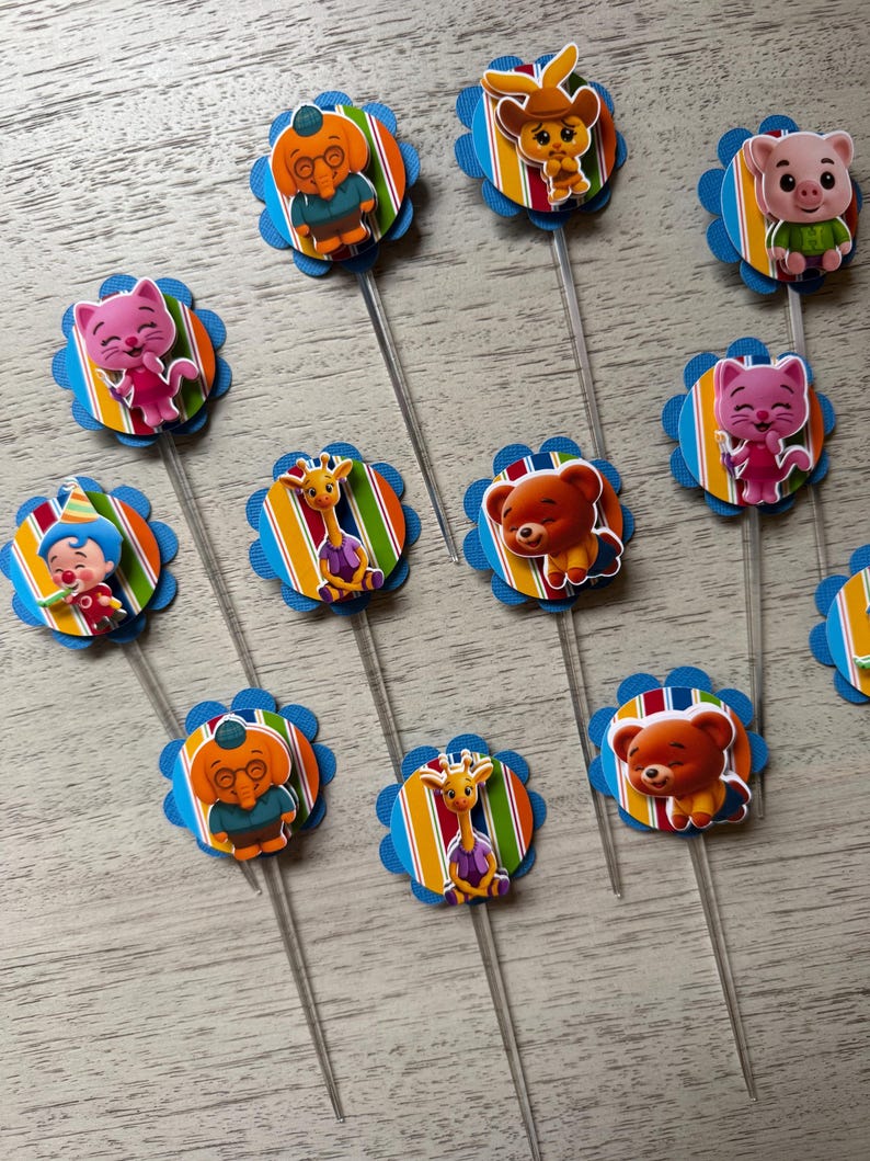 May include: A collection of colorful cupcake toppers featuring various cartoon animal characters. Each topper has a blue scalloped edge, a striped background, and a clear plastic stick.