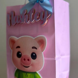 Puede incluir: Bolsa de regalo rosa con una ilustraci&oacute;n de cerdo de dibujos animados con una camisa verde. La bolsa tiene un lazo azul claro y el nombre "Ashley" en una fuente cursiva. Las asas blancas est&aacute;n unidas a la parte superior de la bolsa.