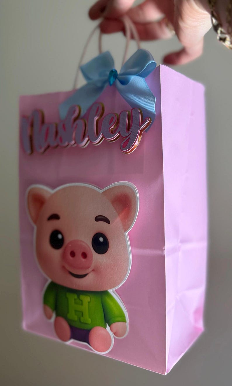 Puede incluir: Bolsa de regalo rosa con una ilustraci&oacute;n de cerdo de dibujos animados con una camisa verde. La bolsa tiene un lazo azul claro y el nombre "Ashley" en una fuente cursiva. Las asas blancas est&aacute;n unidas a la parte superior de la bolsa.