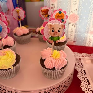 Pu&ograve; includere: Cupcake con glassa rosa e gialla, decorati con personaggi. I cupcake sono esposti su un supporto bianco a pi&ugrave; livelli. Lo sfondo include decorazioni per feste e sacchetti regalo.