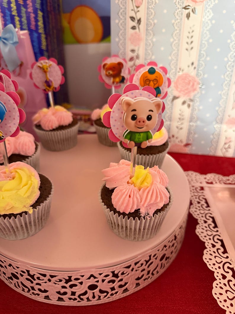 Pu&ograve; includere: Cupcake con glassa rosa e gialla, decorati con personaggi. I cupcake sono esposti su un supporto bianco a pi&ugrave; livelli. Lo sfondo include decorazioni per feste e sacchetti regalo.