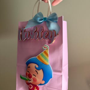 Puede incluir: Bolsa de regalo rosa con asas blancas, un lazo azul claro y el nombre "Ashley" en cursiva. Un personaje de dibujos animados con pelo azul y un gorro de fiesta est&aacute; en la parte delantera, sosteniendo un silbato verde. Una bolsa de fiesta festiva.
