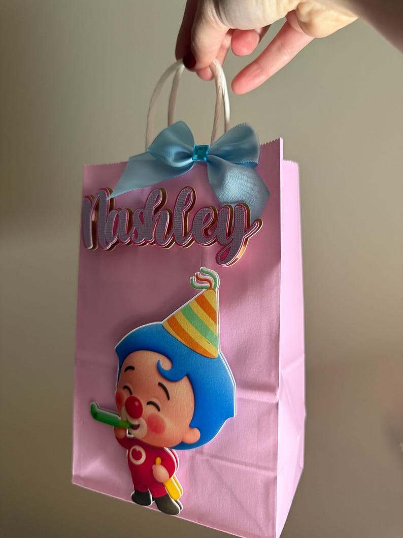 Puede incluir: Bolsa de regalo rosa con asas blancas, un lazo azul claro y el nombre "Ashley" en cursiva. Un personaje de dibujos animados con pelo azul y un gorro de fiesta est&aacute; en la parte delantera, sosteniendo un silbato verde. Una bolsa de fiesta festiva.