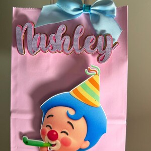 Puede incluir: Bolsa de regalo rosa con un lazo azul claro y el nombre "Nashley" en cursiva. Un personaje de dibujos animados con pelo azul y un gorro de fiesta aparece en la bolsa, sosteniendo un silbato verde.