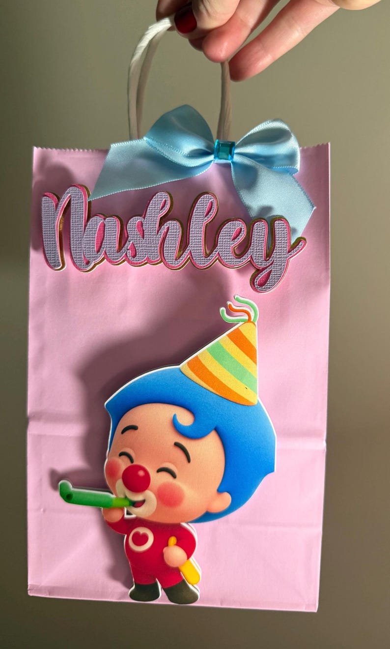 Puede incluir: Bolsa de regalo rosa con un lazo azul claro y el nombre "Nashley" en cursiva. Un personaje de dibujos animados con pelo azul y un gorro de fiesta aparece en la bolsa, sosteniendo un silbato verde.