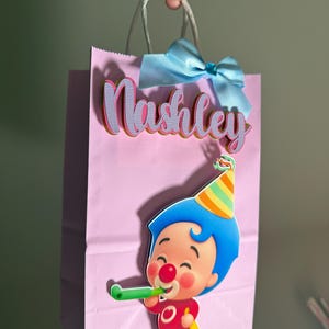 Puede incluir: Una bolsa de regalo rosa con asas de cuerda blanca, un lazo azul claro y el nombre "Nashley" en letras rosas y moradas. Un payaso de dibujos animados con pelo azul y un gorro de fiesta sopla una bocina verde.