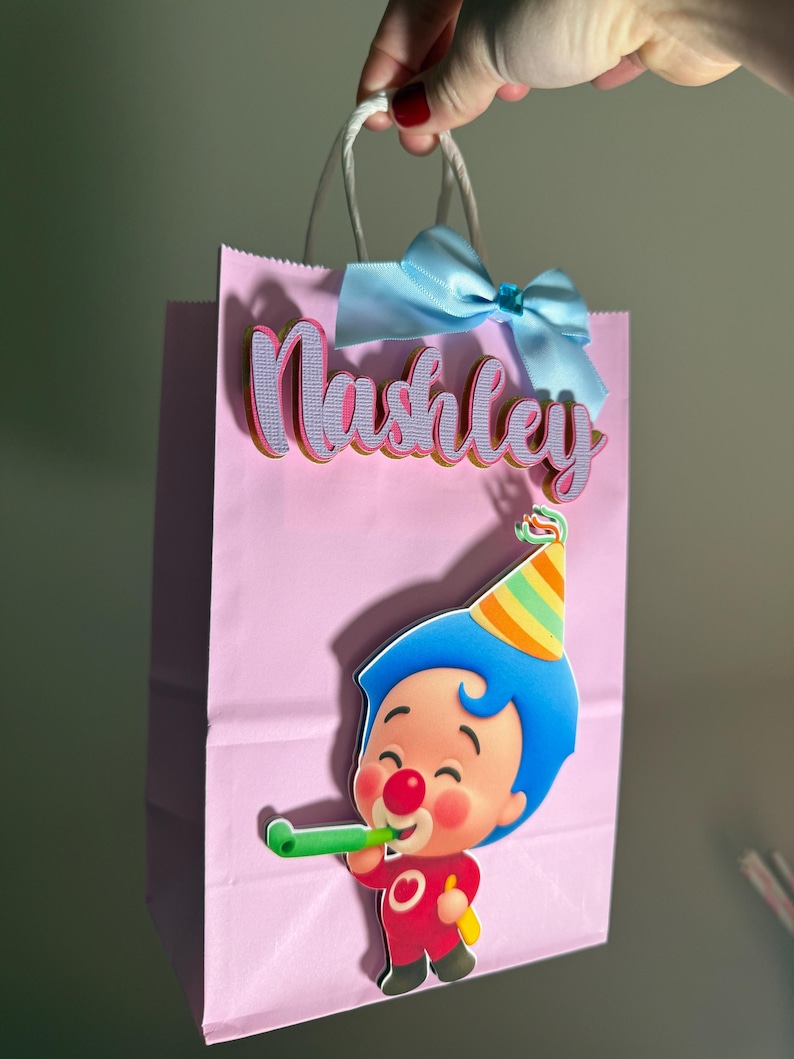 Puede incluir: Una bolsa de regalo rosa con asas de cuerda blanca, un lazo azul claro y el nombre "Nashley" en letras rosas y moradas. Un payaso de dibujos animados con pelo azul y un gorro de fiesta sopla una bocina verde.