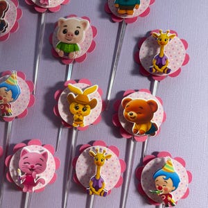 Pu&ograve; includere: Una collezione di decorazioni per cupcake colorate con animali e personaggi dei cartoni animati. Ogni decorazione presenta un'immagine del personaggio su un cerchio rosa a pois, circondato da una forma di fiore rosa, attaccato a un bastoncino di plastica trasparente. I personaggi includono un gatto, un maiale, una giraffa, un orso e un clown.