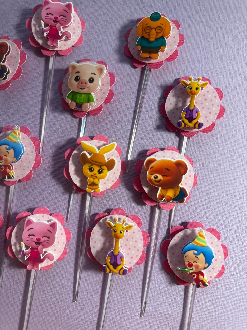 Pu&ograve; includere: Una collezione di decorazioni per cupcake colorate con animali e personaggi dei cartoni animati. Ogni decorazione presenta un'immagine del personaggio su un cerchio rosa a pois, circondato da una forma di fiore rosa, attaccato a un bastoncino di plastica trasparente. I personaggi includono un gatto, un maiale, una giraffa, un orso e un clown.