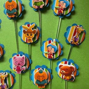 May include: A collection of colorful cupcake toppers featuring cartoon characters. Each topper has a character image on a striped circle with a blue scalloped edge, attached to a clear stick. Characters include a pig, elephant, clown, cat, bear, giraffe, and rabbit.