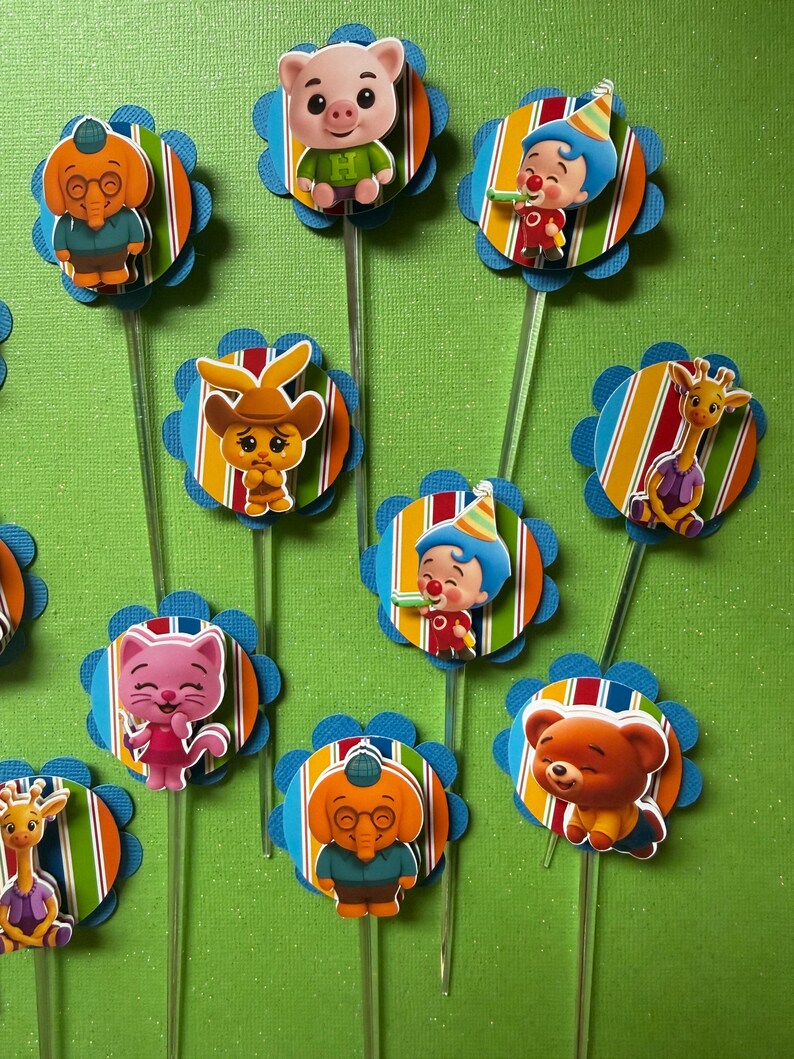 May include: A collection of colorful cupcake toppers featuring cartoon characters. Each topper has a character image on a striped circle with a blue scalloped edge, attached to a clear stick. Characters include a pig, elephant, clown, cat, bear, giraffe, and rabbit.
