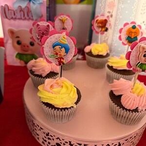 Pu&ograve; includere: Cupcake con glassa rosa e gialla, decorati con personaggi, presentati su un vassoio bianco a pi&ugrave; livelli. Un sacchetto regalo rosa con un'immagine di maiale e il nome "Lushly" &egrave; visibile sullo sfondo.