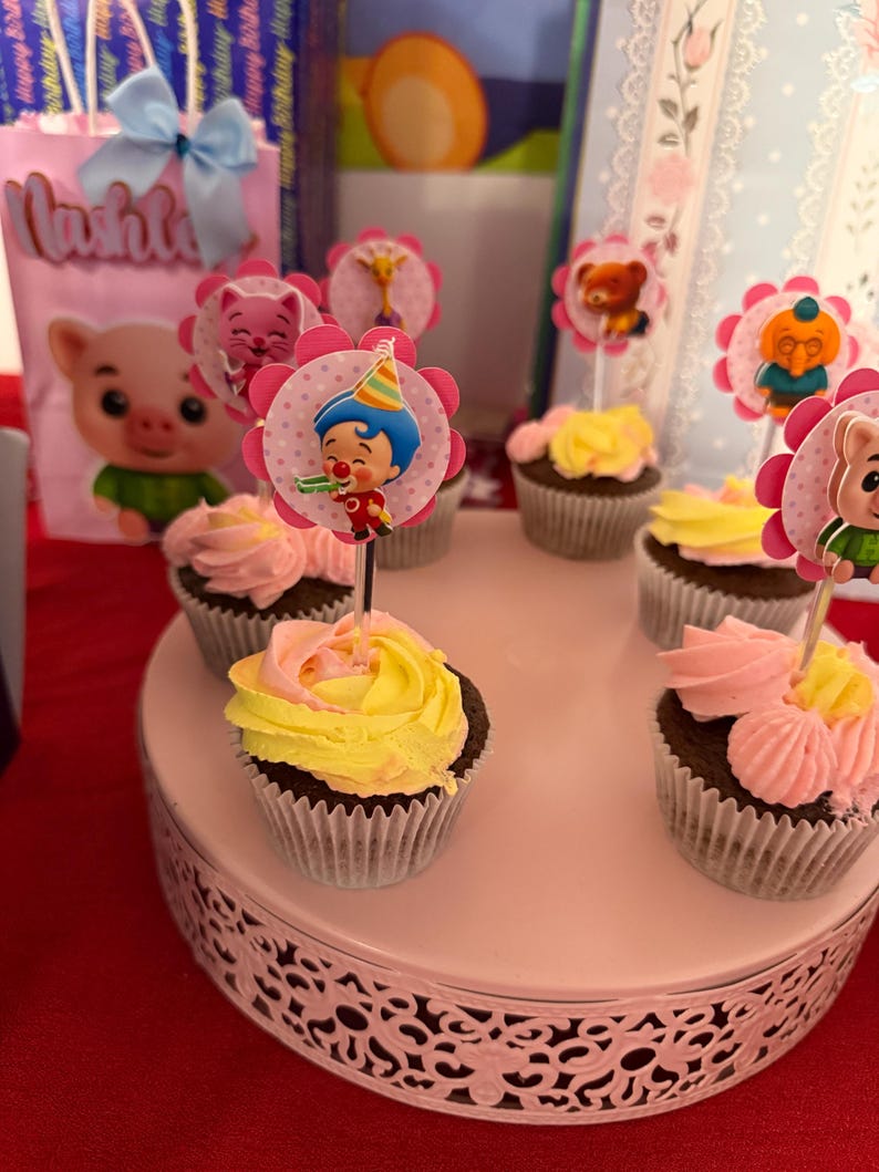 Pu&ograve; includere: Cupcake con glassa rosa e gialla, decorati con personaggi, presentati su un vassoio bianco a pi&ugrave; livelli. Un sacchetto regalo rosa con un'immagine di maiale e il nome "Lushly" &egrave; visibile sullo sfondo.