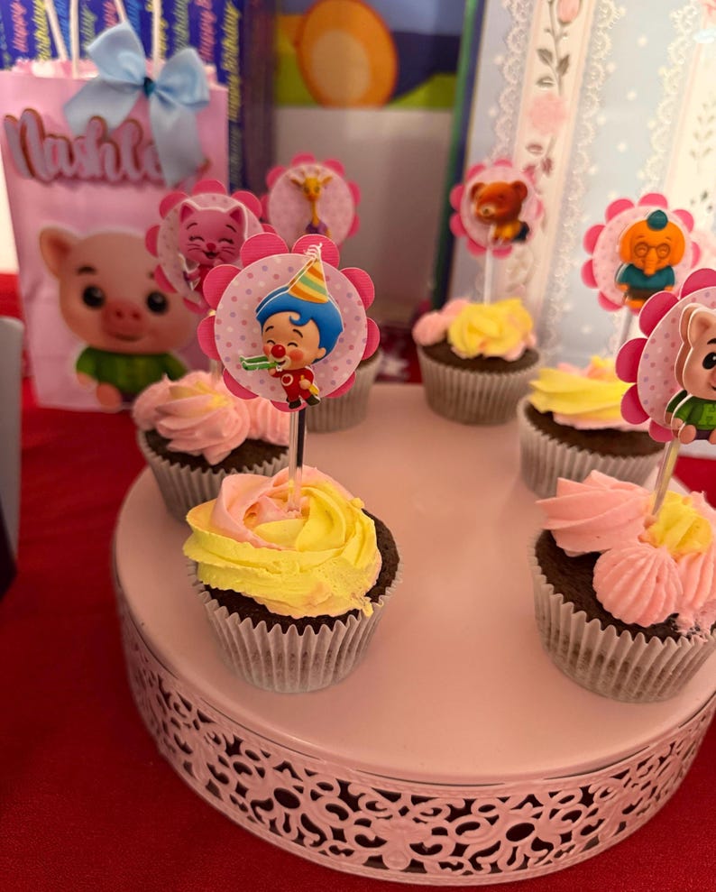 Pu&ograve; includere: Cupcake con glassa rosa e gialla, decorati con topper a forma di personaggi. I cupcake sono disposti su un alzatina rosa e decorativa. Sullo sfondo, una borsa regalo rosa con l'illustrazione di un maiale.