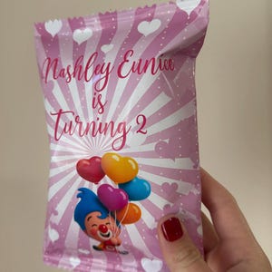 May include: Pink party favor bag with white hearts and stripes. Text reads "Nashley Eunice is Turning 2". A cartoon clown holds heart-shaped balloons in red, orange, yellow, purple, and blue. The bag is held by a person with red nail polish.