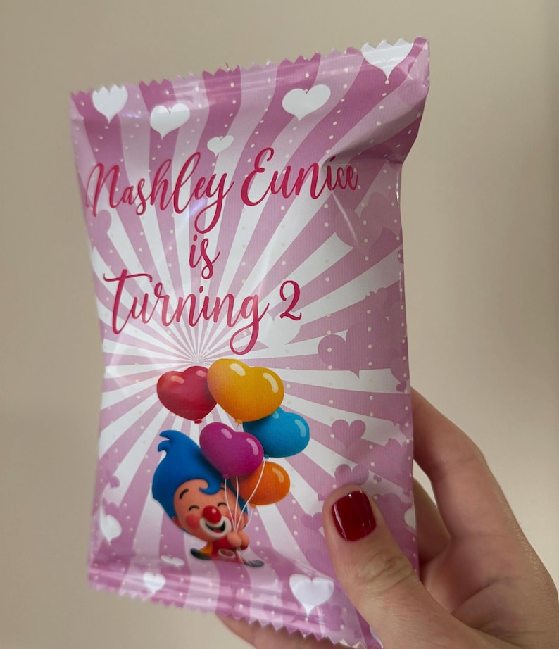 May include: Pink party favor bag with white hearts and stripes. Text reads "Nashley Eunice is Turning 2". A cartoon clown holds heart-shaped balloons in red, orange, yellow, purple, and blue. The bag is held by a person with red nail polish.