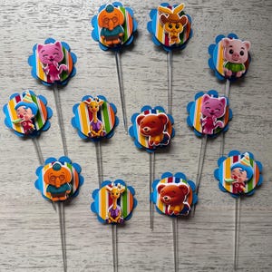 May include: A collection of colorful cupcake toppers featuring various cartoon animal characters. Each topper has a blue scalloped edge, a striped background, and a clear plastic stick.