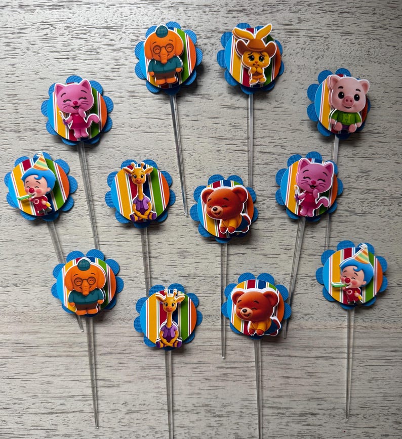 May include: A collection of colorful cupcake toppers featuring various cartoon animal characters. Each topper has a blue scalloped edge, a striped background, and a clear plastic stick.
