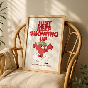 May include: A framed print with the text "JUST KEEP SHOWING UP" in red, above a cartoon strawberry character. The strawberry has arms, legs, and a stern expression. The text "The accolades will follow" is below the strawberry.