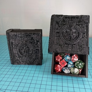 May include: Two black, book-shaped dice boxes with intricate relief carvings. One box is closed, the other open, revealing a collection of colorful polyhedral dice. The boxes are on a blue grid surface.