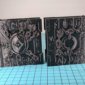 May include: Two black, rectangular bookends with a detailed, embossed design. Each bookend features a central shield or circular element, surrounded by chains and decorative embellishments. The bookends are designed to resemble ancient, weathered stone.