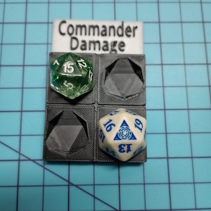 May include: A 3D printed holder for dice, labeled "Commander Damage". The holder has four slots, two of which are filled with dice. One die is green with white numbers, the other is white with blue numbers and a blue symbol.