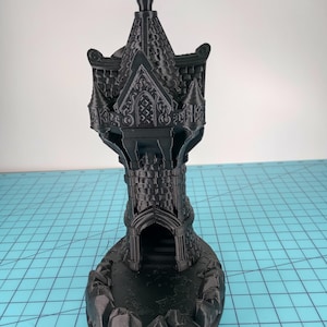 May include: A black, 3D-printed castle tower with intricate architectural details. The tower features a pointed roof, arched doorways, and decorative elements. The base has a rocky terrain design. The overall height is approximately 6 inches.