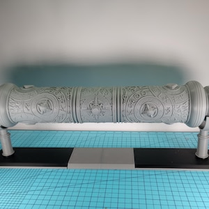 May include: A gray, cylindrical decorative object with intricate engraved patterns, supported by a black and gray base. The object features detailed circular and star-like designs. The base has two vertical supports. The overall design suggests a historical or fantasy theme.
