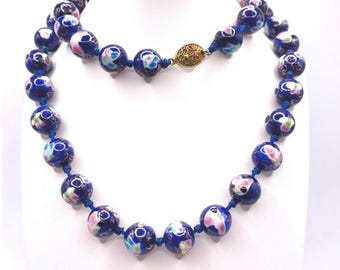 Könnte beinhalten: Eine blaue Perlenkette mit floralen Designs in Rosa, Weiß und Grün. Die Halskette hat runde Perlen, die auf einer blauen Schnur aufgereiht sind, mit einem goldfarbenen Verschluss. Die Perlen haben ein glänzendes Finish.