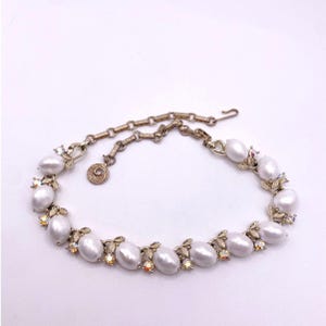 Puede incluir: Un collar vintage con grandes perlas de imitación ovaladas. El collar tiene detalles metálicos dorados con adornos de hojas y cristales. El cierre y la cadena también son dorados, creando un diseño clásico y elegante.