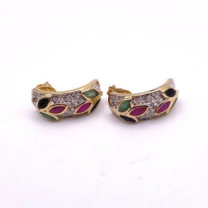 May include: A pair of gold-toned hoop earrings. Each earring features a curved design with a row of small, clear stones and colorful leaf-shaped accents in black, green, and red. The earrings are set against a white background.