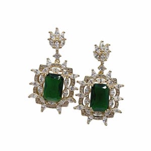 Puede incluir: Un par de pendientes dorados con piedras centrales rectangulares de color verde esmeralda. Los pendientes tienen un halo de piedras claras y brillantes y una parte superior decorativa con más piedras claras. Los pendientes están sobre un fondo blanco.